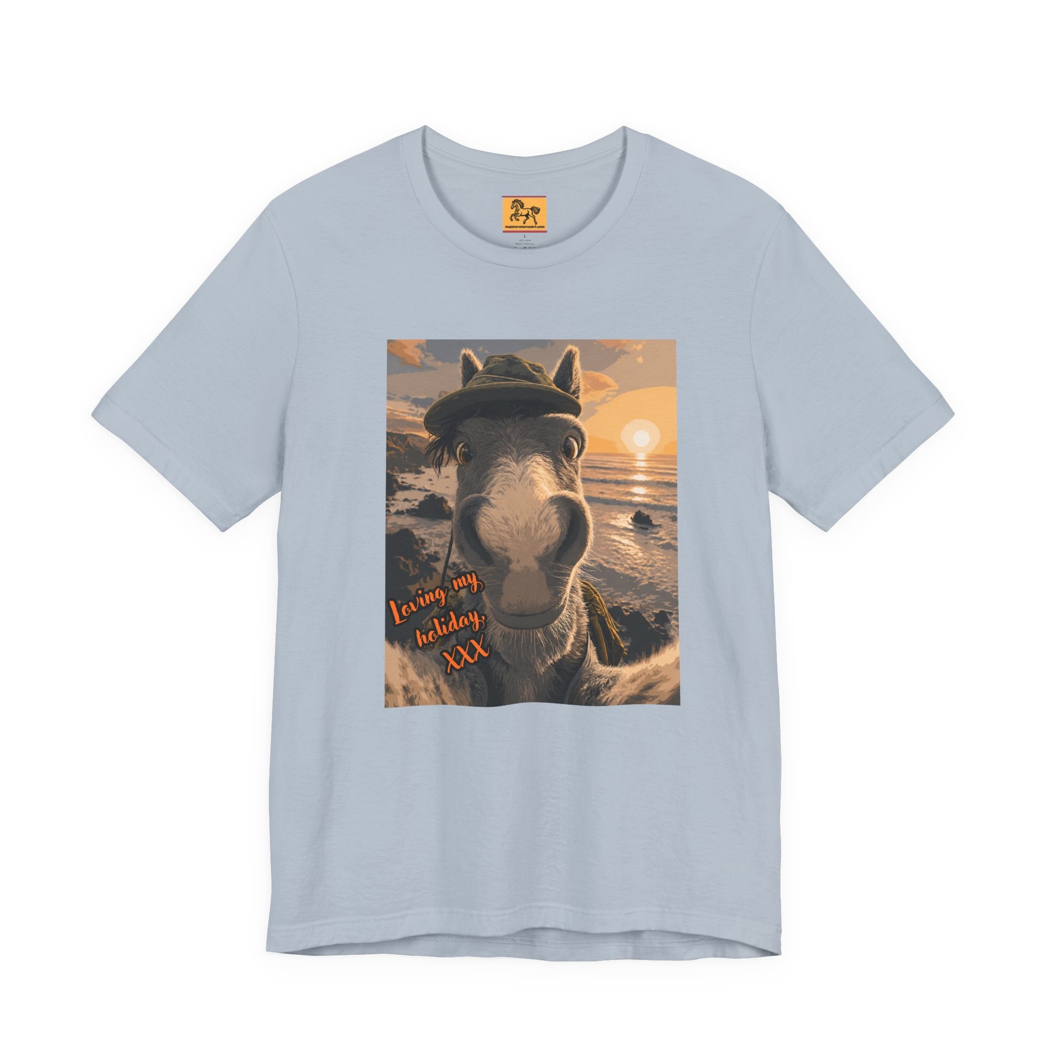A light blue Bella + Canvas 3001 unisex t-shirt featuring an illustrated horse taking a beach selfie at sunset, wearing a hat, with the words 'Loving my holiday XXX' in orange script. 