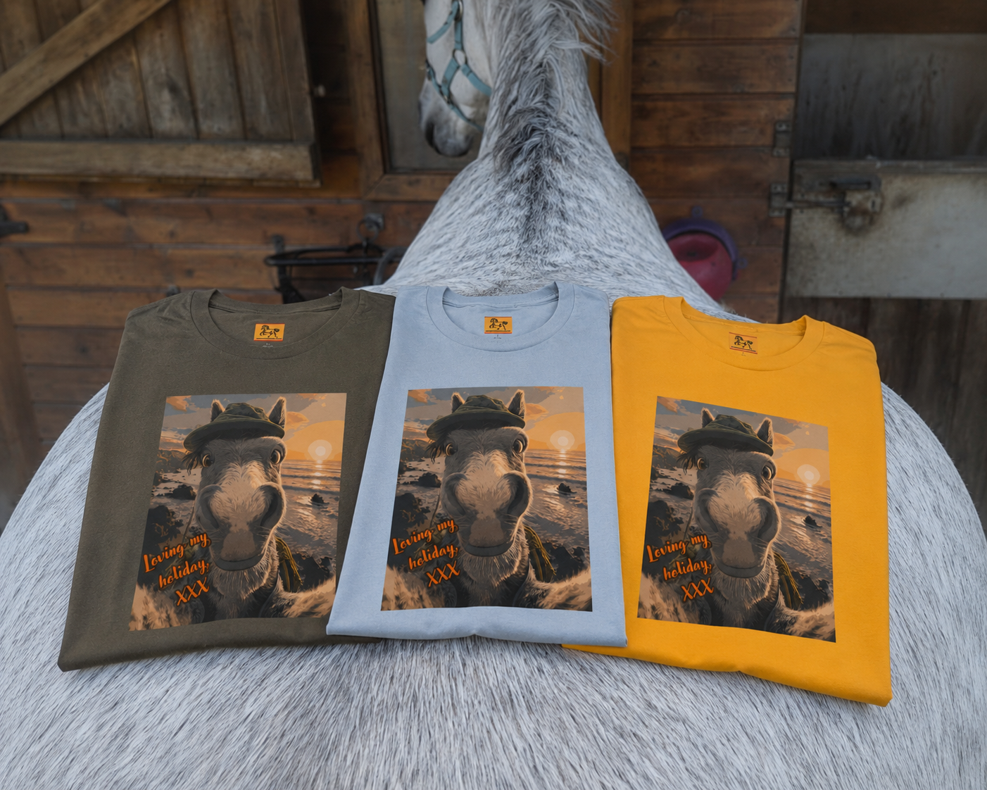 Three folded Bella + Canvas 3001 unisex t-shirt featuring an illustrated horse taking a beach selfie at sunset, wearing a hat, with the words 'Loving my holiday XXX' in orange script. Resting on a horses back.